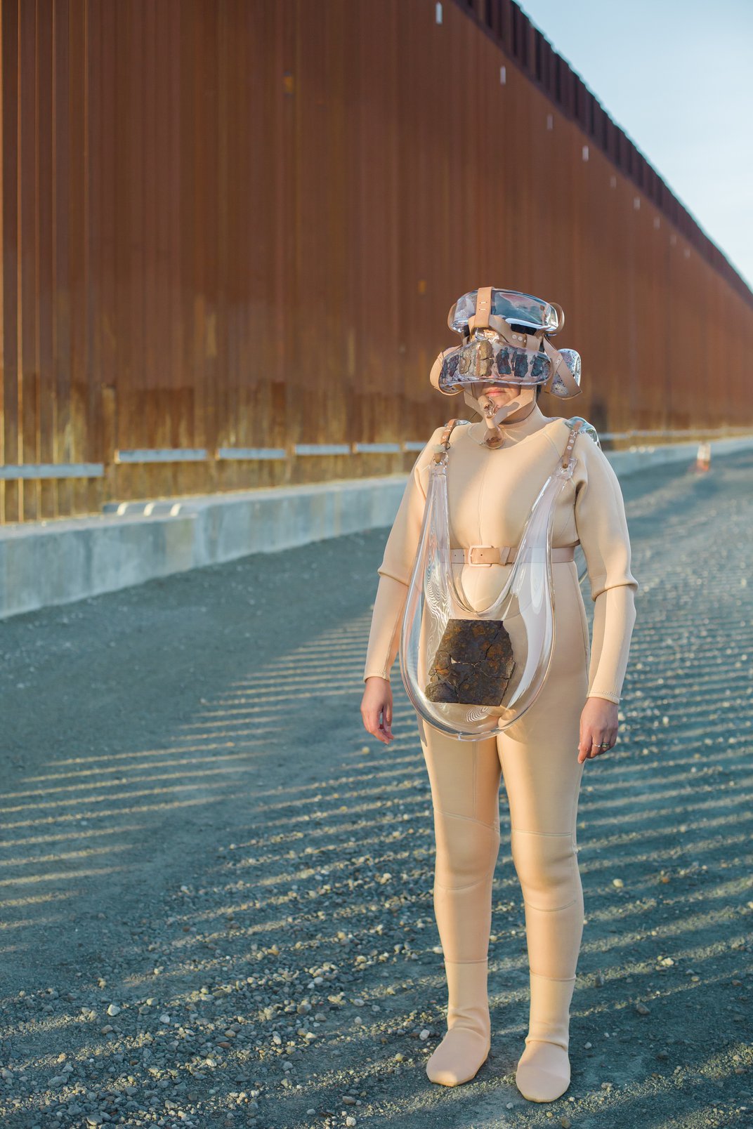 Artist Tanya Aguiniga standing in a suit made of various glass elements next to the U.S./ Mexico border wall.